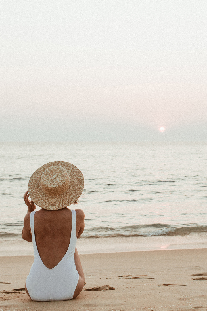 woman on beach wearing white swimsuit and sun hat watching the sunset